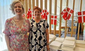 Tine Hansen og Anne Sofie, står foran de store bunker af trykte begynderbøger nr. 1-5. og med flag i baggrunden.