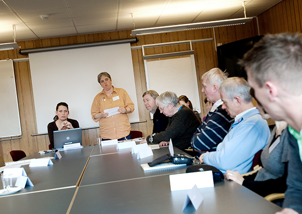 Birgit Christensen står for enden af et bord og holder oplæg. Personer rundt om bordet lytter.