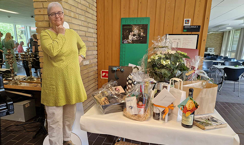 En papfigur af Birgit foran gavebordet fra receptionen, fyldt med blomster og vin m.m.